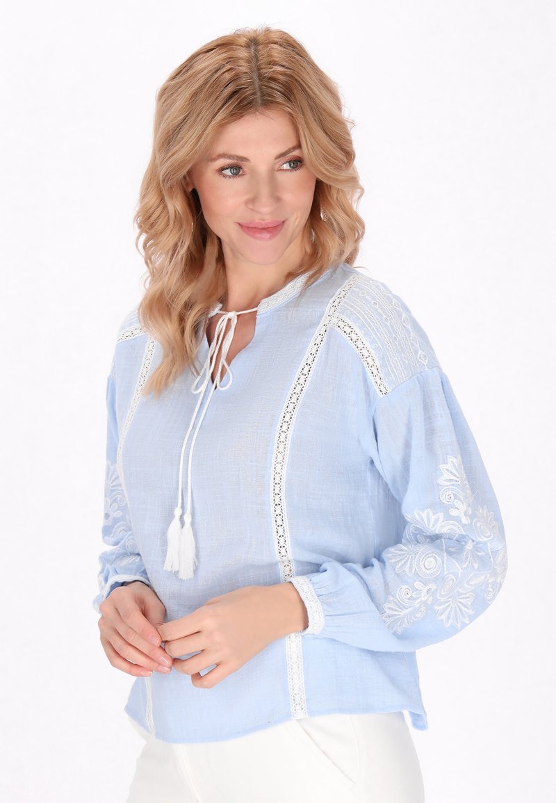 Blusa azzurro chiaro realizzata in tessuto testurizzato, con dettagli in pizzo bianco e ricami floreali sulle maniche. Si lega con dei ciondoli sul collo.