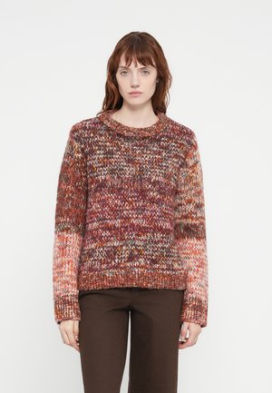 Knit sweater in warm hues of red, orange, and cream with a loose fit, crew neckline, and textured pattern. Paired with brown pants.