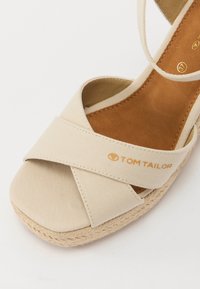 Beige lerretsandal med kryssede reimer, en polstret brun såle og en flettet jutesåle med en detalj i gull.