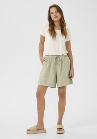 Šviesiai smėlinis marškinėliai derinami su šviesiai žaliais, iki kelių siekiančiais šortais, turinčiais raištelio juosmenį ir didelėmis priekinėmis kišenėmis. Dėvimi su smėlio spalvos šlepetėmis.