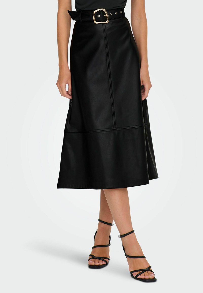 Femme portant une jupe midi en cuir noire taille haute avec une large ceinture et des sandales à talons avec des brides noires, se tenant les jambes croisées.