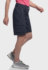 Marinefarbene Cargo-Shorts aus leichtem Stoff. Mit einer vorderen Reißverschlusstasche und einer großen Seitentasche ausgestattet. Kombiniert mit grauen Sportschuhen.