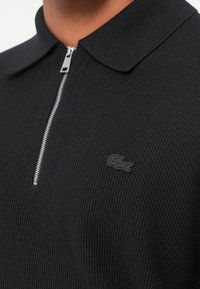 Schwarzes Poloshirt aus strukturiertem Stoff, mit einem silbernen Reißverschluss und einem kleinen schwarzen Logo auf der Brust.