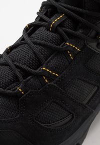 Baskets noires en tissu texturé et empiècements en daim, avec des lacets noirs passés à travers des boucles ornées de coutures jaunes.