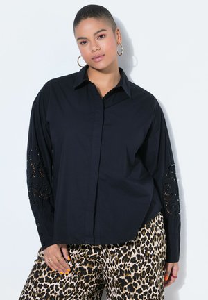 Frau mit kurzen Haaren, die ein schwarzes Hemd mit Spitzenärmeln und eine Hose mit Leopardenmuster trägt, steht vor einem weißen Hintergrund.