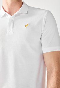 White polo shirt made of textured fabric, featuring a flat collar, short sleeves, and a small golden eagle logo on the left chest.