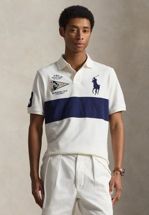 Homme portant un polo blanc avec une bande bleu marine, un logo joueur de polo bleu marine et un pantalon blanc plissé, debout devant un fond uni.