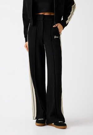 Personne portant un pantalon de jogging noir à jambes larges avec des bandes latérales beige, un petit logo "Guess", associé à des baskets noires à semelles en gomme.