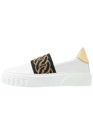 Valkoinen slip-on-tennari, jossa on musta hihna kullanvärisillä strassikoristeilla, kultaista tehostetta kantapäässä ja teksturoitu kumipohja.