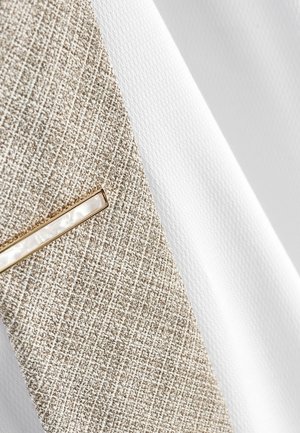 Tessuto beige chiaro con trama a griglia e una sezione bianca, caratterizzato da un dettaglio dorato con un particolare simile a una perla.