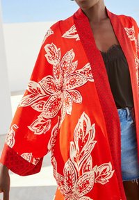 Kimono rouge avec de grandes impressions florales blanches et des accents à motif de treillis. Tissu léger, coupe ample, manches larges et ourlet atteignant les hanches.
