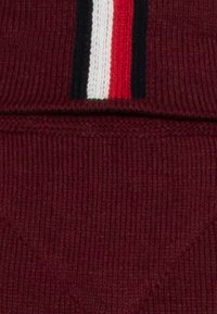Burgundy stickad tyg med ett horisontellt randmönster i röd, vit och svart färg nära den övre kanten. Mjuk och elastisk textur.