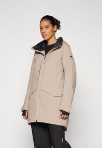 Mujer que lleva una parka beige con capucha y forro de forro polar, manga larga negra debajo y pantalones negros con un logo en la pierna izquierda.