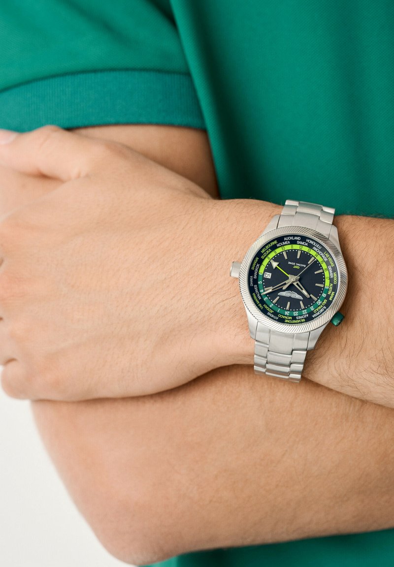 Osoba nosząca srebrny zegarek na rękę z czarno-zielonym cyferblatem światowego czasu, mająca skrzyżowane ramiona w zielonej koszulce z krótkimi rękawami.