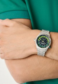 Persona che indossa un orologio da polso argento con un quadrante mondiale nero e verde, con le braccia incrociate in una camicia verde a maniche corte.