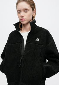 Jachetă din fleece neagră, cu guler înalt, fermoar frontal și detaliu cu logo. Material texturat cu un aspect moale și pufos. Dispune de două buzunare frontale.
