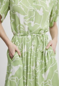 Rochie verde cu un model abstract alb, maneci scurte, închidere cu nasturi în față, talie strânsă și buzunare laterale. Material moale cu o textură fluidă.