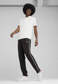 Hvid polo-shirt med Ferrari-logo, sorte sweatpants med sidelæg, og hvide sneakers med røde accenter. Modellen står i en afslappet position.