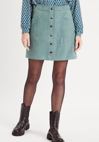 Jupe mini en daim vert avec des boutons sur le devant, deux poches latérales et des coutures plates. Portée avec des bottines noires et des collants noirs transparents.