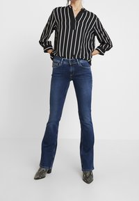 Gestreifte Bluse in Schwarz-Weiß mit Kragen, kombiniert mit dunkelblauen Schlagjeans und gemusterten ankle boots. Einheitlicher weißer Hintergrund.