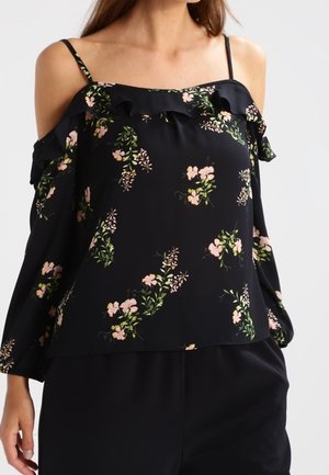 Femme portant un chemisier noir à épaules dénudées avec imprimé floral rose et vert et un col à volants, associé à un pantalon noir.