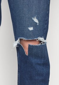 Jeans de mezclilla azul con desgastes rasgados y bordes deshilachados. La tela tiene un tejido clásico de mezclilla con una textura suave y un sutil desgaste.