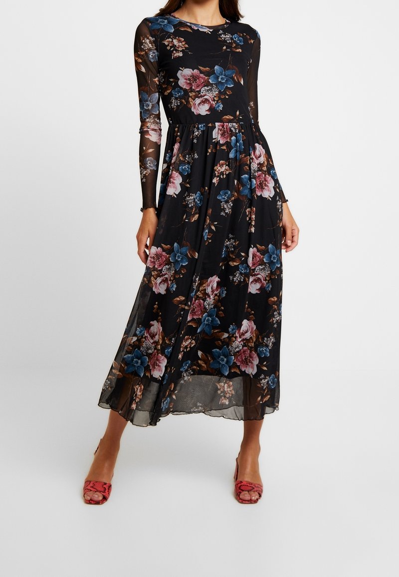 Femme portant une robe noire transparente à manches longues avec des motifs floraux roses et bleus et des sandales à talons rouges.