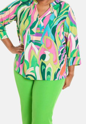 Osoba ubrana w jaskrawozielone spodnie i bluzkę z abstrakcyjnym, wielokolorowym wzorem w odcieniach zieleni, różu, niebieskiego i fioletu.