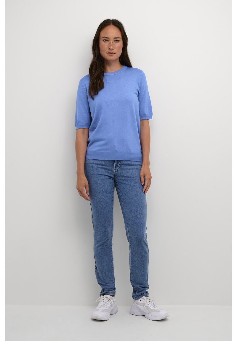 Frau mit langen dunklen Haaren, die einen blauen Kurzarmpullover, blaue Jeans und weiße Turnschuhe trägt und vor einem schlichten hellen Hintergrund steht.