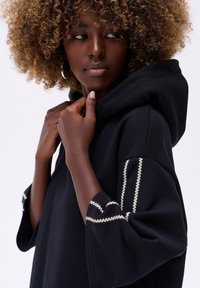 Czarna bluza z kapturem o luźnym kroju. Wyróżnia się dekoracyjnym białym przeszyciem na mankietach. Miękka faktura i regulowany kaptur.