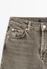 Jeans in denim grigio con una leggera lucentezza, dotati di un design classico a cinque tasche, chiusura con bottone in metallo e dettagli cuciti. Etichetta del marchio visibile.