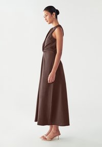 Vestido marrón sin mangas con diseño de bodice torcido, hasta el suelo, hecho de tela suave. Modelo con sandalias de tacón nude.