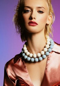 Young woman with short blonde hair wearing layered large blue and white bead necklaces and a shiny pink satin blouse against purple background.