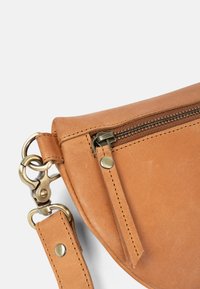 Eck eines braunen Lederbeutels mit Messing-Reißverschluss und -Verschluss, angenähtem Riemen, der an Metallringen auf weißem Hintergrund befestigt ist.