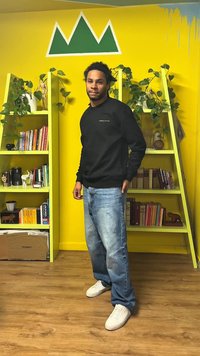 Mies mustassa collegepaidassa ja farkuissa seisoo kahden keltaisen kirjahyllyn välissä, joissa on kasveja ja kirjoja, puisella lattialla vihreän vuorimurataustakuvan alla.