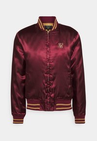 Bordeaux satin bomberjakke med ribbet krave og manchetter, som har guldaccentstriber og et lille guldlogo på brystet.