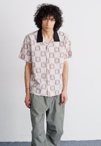 Short-sleeve button-up shirt in cream with maroon geometric patterns, featuring a black collar. Worn with loose-fitting gray pants.