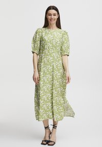Robe midi fleurie verte avec des manches bouffantes, ornée de motifs de fleurs blanches et d'une taille cintrée. Associée à des talons strappy noirs.
