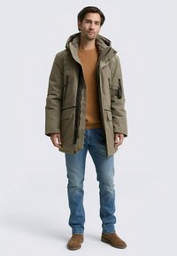 Olivzaļš parka mētelis ar kapuci, vairākām kabatām un rāvējslēdzēja priekšpusi. Valkāts pār brūnu džemperi un zilas džinsas, kopā ar brūnām zābakiem.