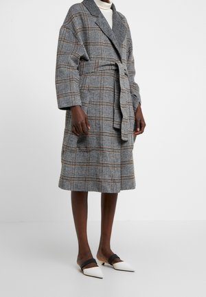 Femme portant un manteau gris à carreaux longueur genoux avec un large col et une ceinture, associé à des mules blanches à bout pointu avec des brides noires.