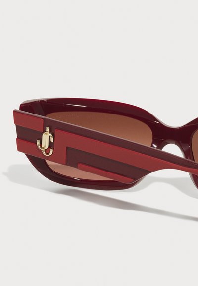 Red sunglasses with a glossy finish, featuring bold geometric accents on the arms and a gold logo detail. Brown tinted lenses complete the design.