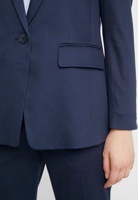 Blazer bleu marine ajusté avec poche à rabat et un bouton, porté avec un pantalon bleu marine assorti, visible de la taille jusqu’au milieu de la cuisse.