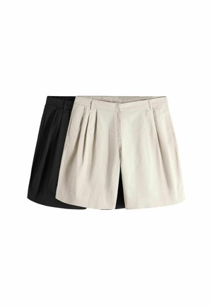 Žena stojaca pri bielej stene, oblečená v tmavohnedom tielku bez rukávov, voľných čiernych nohaviciach nad kolená a čiernych nazúvacích topánkach.