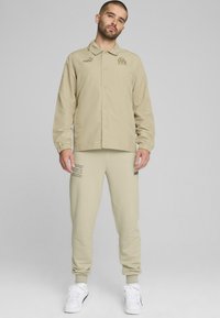 Beige vindjakke med trykknaplukning, rund krave og elastiske manchetter, parret med matchende joggingbukser med logo og dekorative striber.