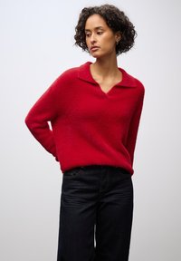 Roter, flauschiger Pullover mit V-Ausschnitt und Kragen-Detail. Kombiniert mit dunkelblauen Jeans im klassischen Schnitt und Nähten.