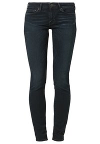 Jean skinny bleu foncé avec fermeture à bouton et fermeture éclair, poches avant et arrière, et légère décoloration sur les cuisses et les genoux.