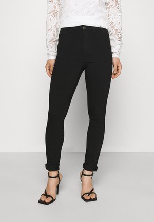 Femme portant un pantalon noir ajusté avec les ourlets retroussés, associé à des sandales noires à talons hauts et bretelles, et un haut à manches longues en dentelle blanche.