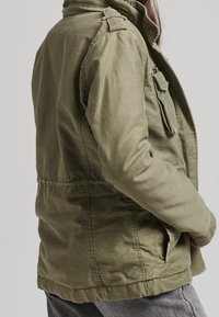 Olivgrön jacka med en texturerad finish, med dragkedja framtill, sidofickor och en figursydd midja. Innehåller axelpartier för detaljer.