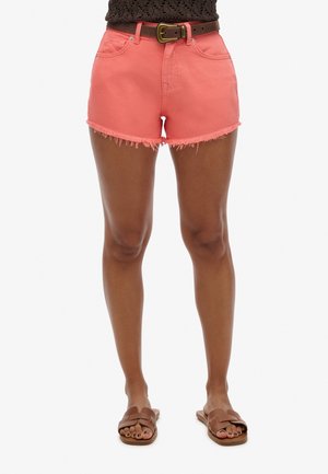 Vrouw die koraalrode shorts met rafelige zoom, een bruine riem, een bruin gehaakt topje en bruine slippertjes draagt, tegen een witte achtergrond.