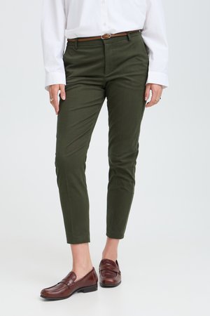 Personne portant un pantalon slim vert olive, une ceinture marron, une chemise blanche boutonnée et des mocassins en cuir marron, debout sur un fond clair.